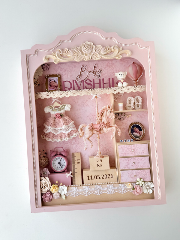 Pink vintage-style nursery shadow box with a carousel horse figure, crocheted dress, miniature dresser drawers, teddy bear, and birth-weight and date blocks