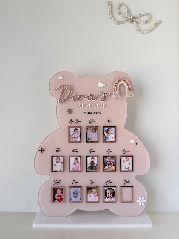 Pink bear-shaped acrylic milestone stand with twelve monthly photo slots and a macrame rainbow charm, tracking a baby's first year
