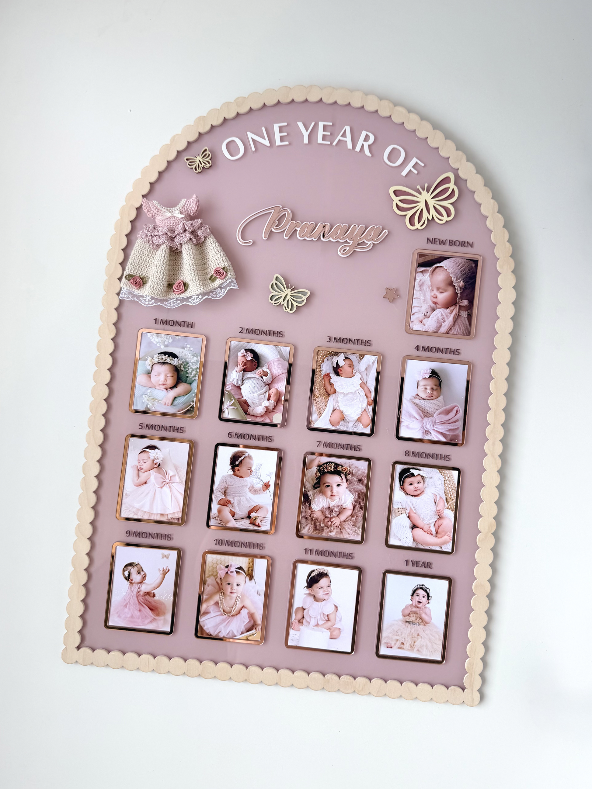 Mauve arch-shaped acrylic milestone board with scalloped wood trim, thirteen photo frames, a crocheted dress, and butterfly accents