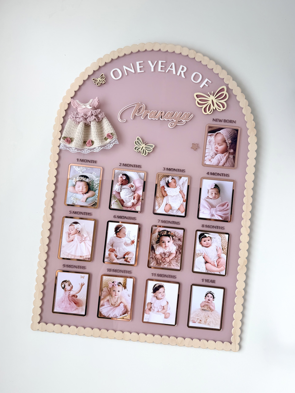 Mauve arch-shaped acrylic milestone board with scalloped wood trim, thirteen photo frames, a crocheted dress, and butterfly accents