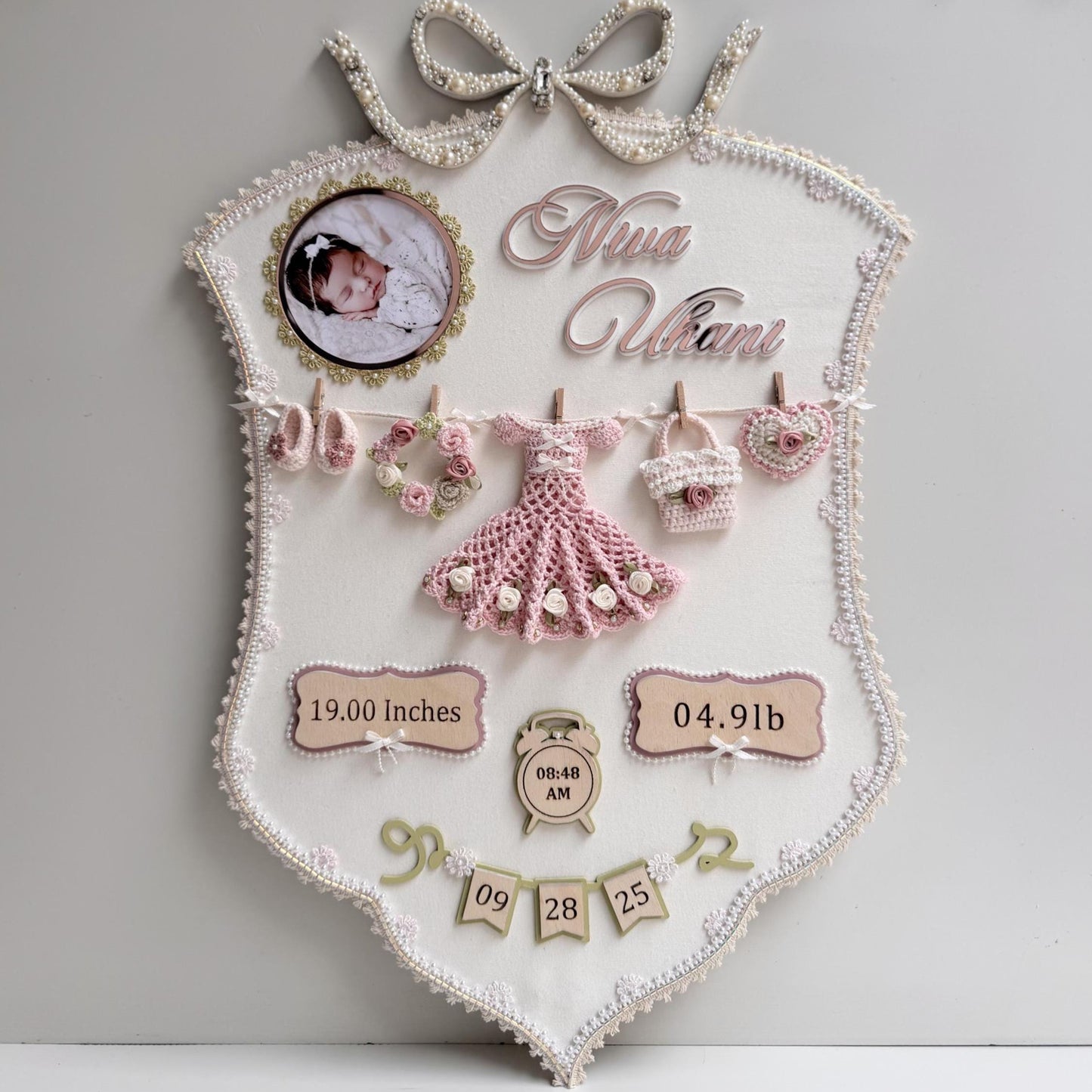 Cream fabric birth-stats keepsake board with a pearl bow, baby photo, and a crocheted dress, wreath, bag, and heart clipped to a mini clothesline