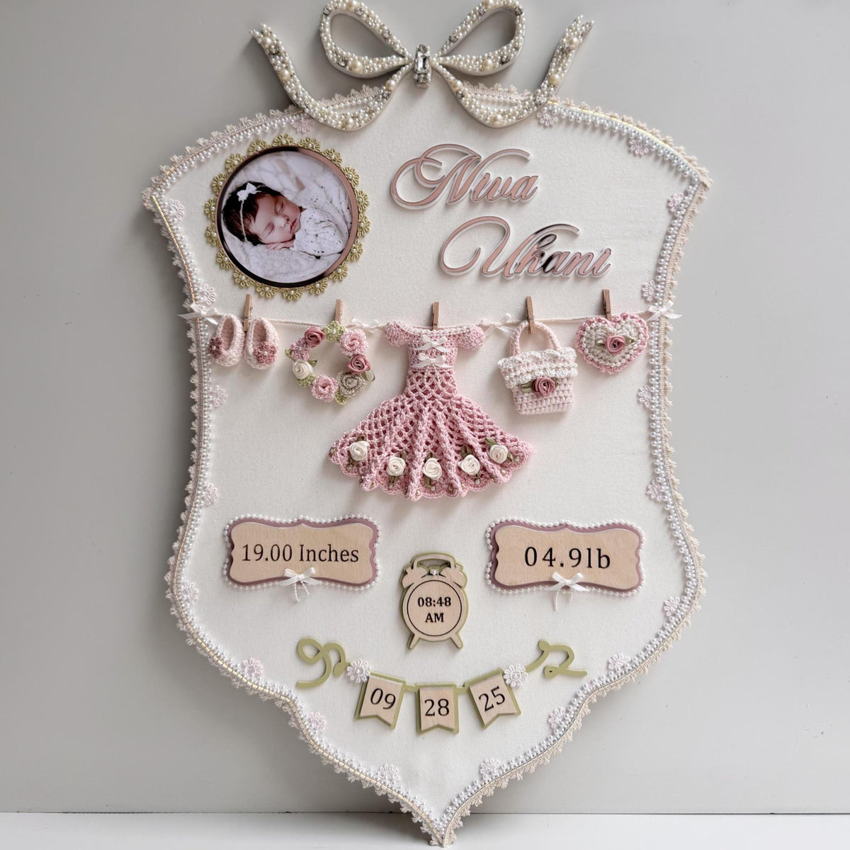 Cream fabric birth-stats keepsake board with a pearl bow, baby photo, and a crocheted dress, wreath, bag, and heart clipped to a mini clothesline