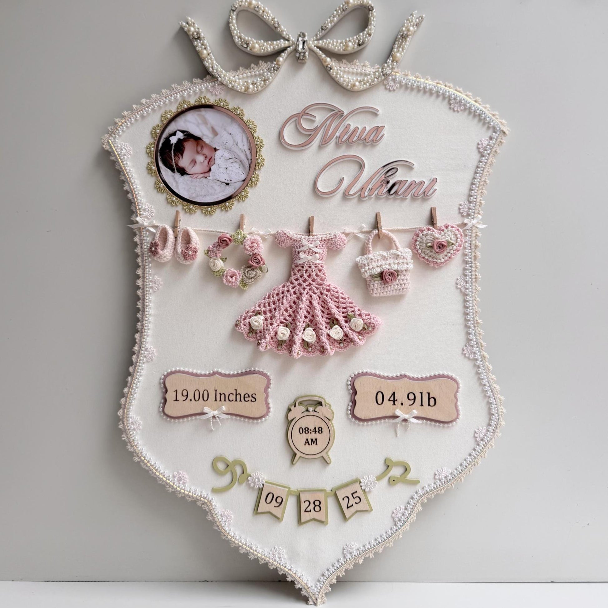 Cream fabric birth-stats keepsake board with a pearl bow, baby photo, and a crocheted dress, wreath, bag, and heart clipped to a mini clothesline