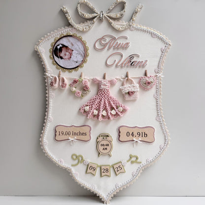 Cream fabric birth-stats keepsake board with a pearl bow, baby photo, and a crocheted dress, wreath, bag, and heart clipped to a mini clothesline