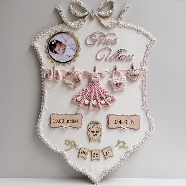 Cream fabric birth-stats keepsake board with a pearl bow, baby photo, and a crocheted dress, wreath, bag, and heart clipped to a mini clothesline