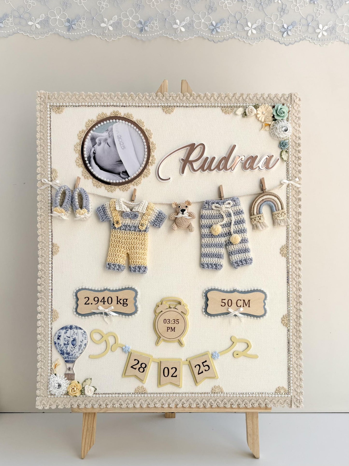 Cream fabric nursery keepsake board on a small easel with a baby photo, the baby's name, a crocheted outfit and booties on a mini clothesline, and birth stats