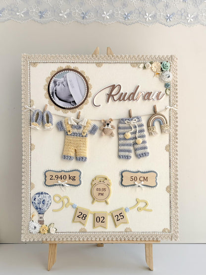 Cream fabric nursery keepsake board on a small easel with a baby photo, the baby's name, a crocheted outfit and booties on a mini clothesline, and birth stats