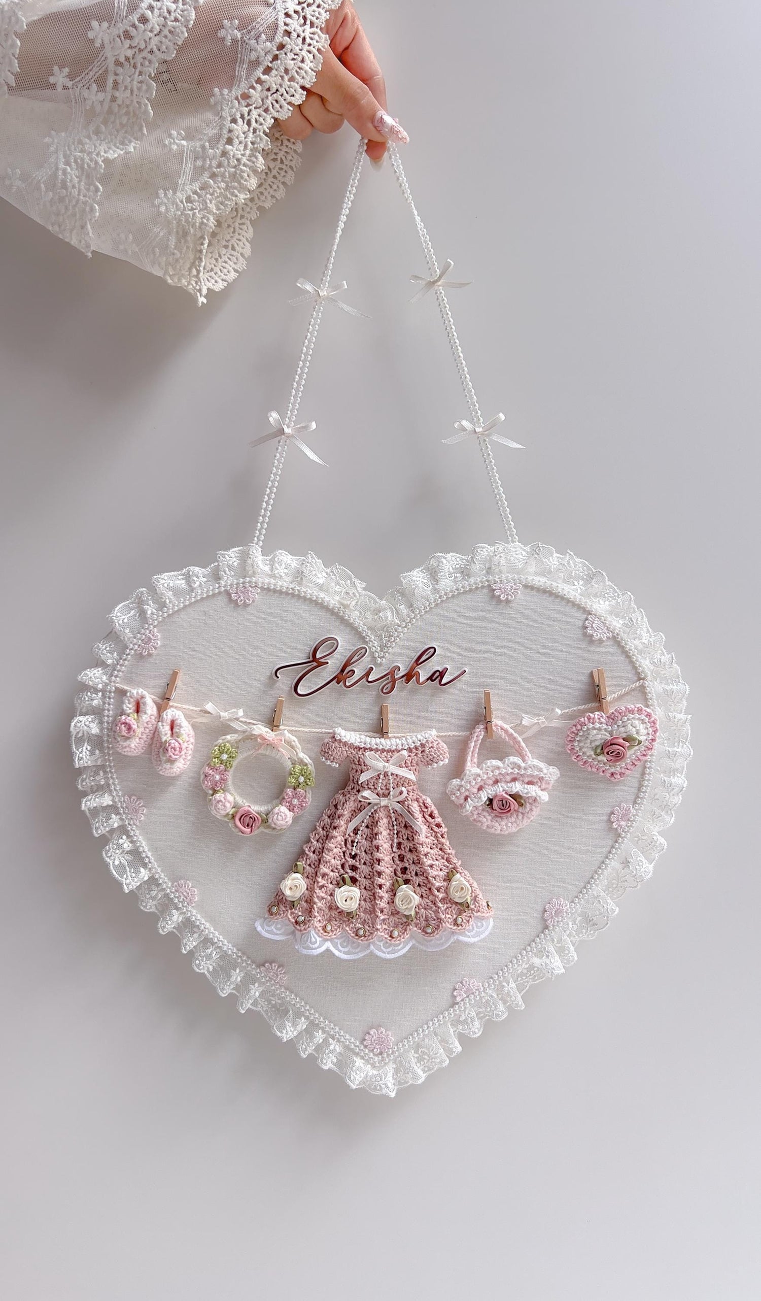 Heart-shaped lace-trimmed fabric wall hanging with the baby's name and a crocheted dress, wreath, basket, and heart clipped to a mini clothesline
