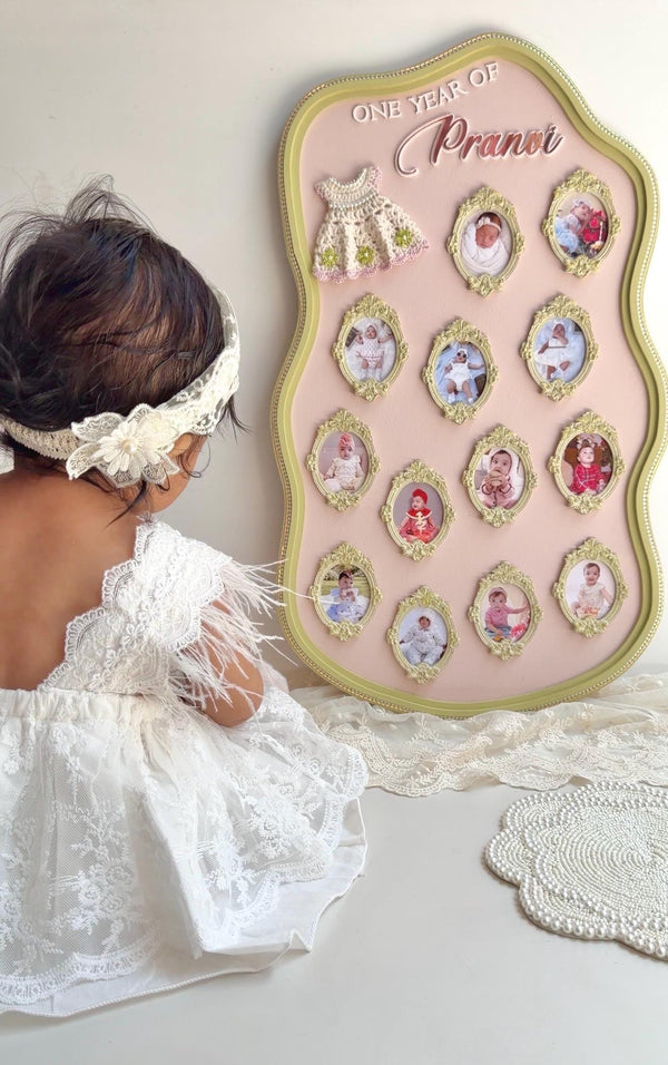 Scalloped pink and gold nursery board with thirteen ornate oval photo frames tracking a baby's first year and a crocheted dress topper