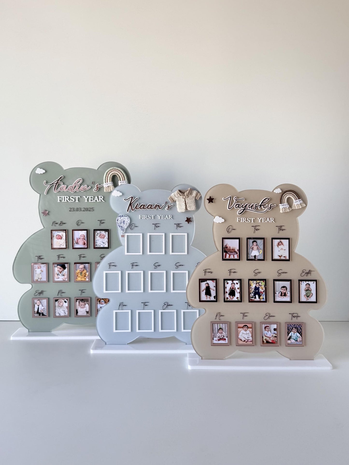 Three bear-shaped acrylic milestone boards in sage, dusty blue, and taupe, each with twelve monthly photo slots and a mini rainbow or sweater charm