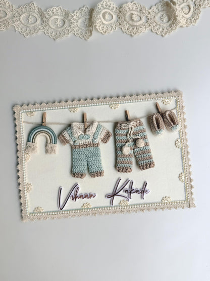 Rectangular fabric keepsake frame with a mini crocheted onesie, pants, and booties clipped to a clothesline, a rainbow charm, and the baby's name