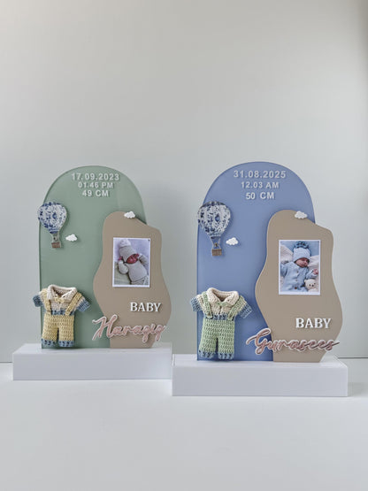 Two arch-shaped acrylic birth-announcement plaques, each with a baby photo, hot-air-balloon charm, crocheted romper, and birth date, time, and length