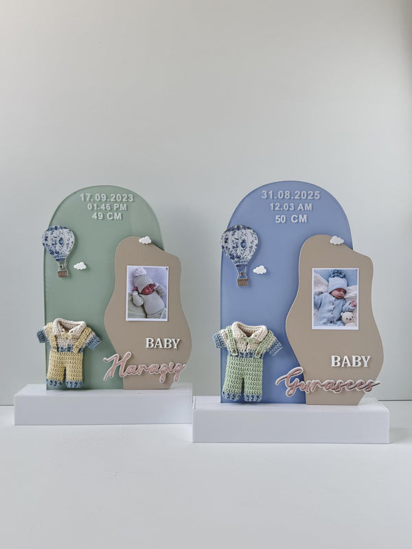 Two arch-shaped acrylic birth-announcement plaques, each with a baby photo, hot-air-balloon charm, crocheted romper, and birth date, time, and length
