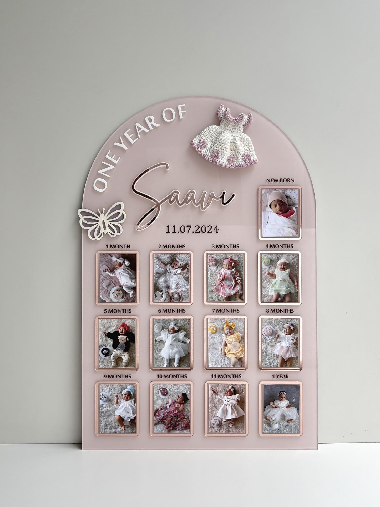 Arch-shaped pink acrylic milestone board with thirteen photo frames from newborn to one year and a crocheted dress topper