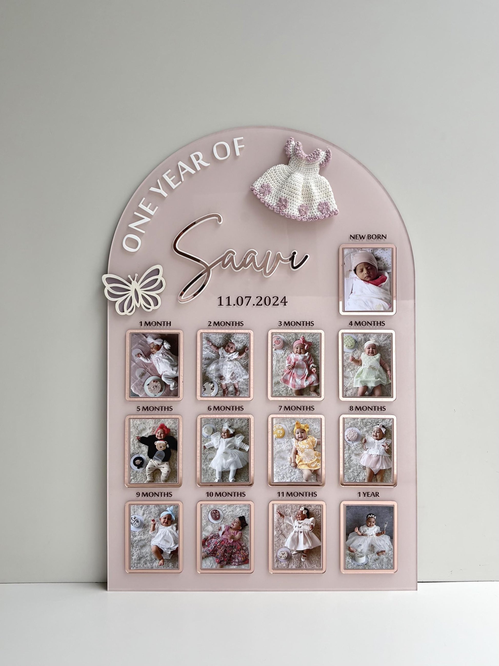 Arch-shaped pink acrylic milestone board with thirteen photo frames from newborn to one year and a crocheted dress topper