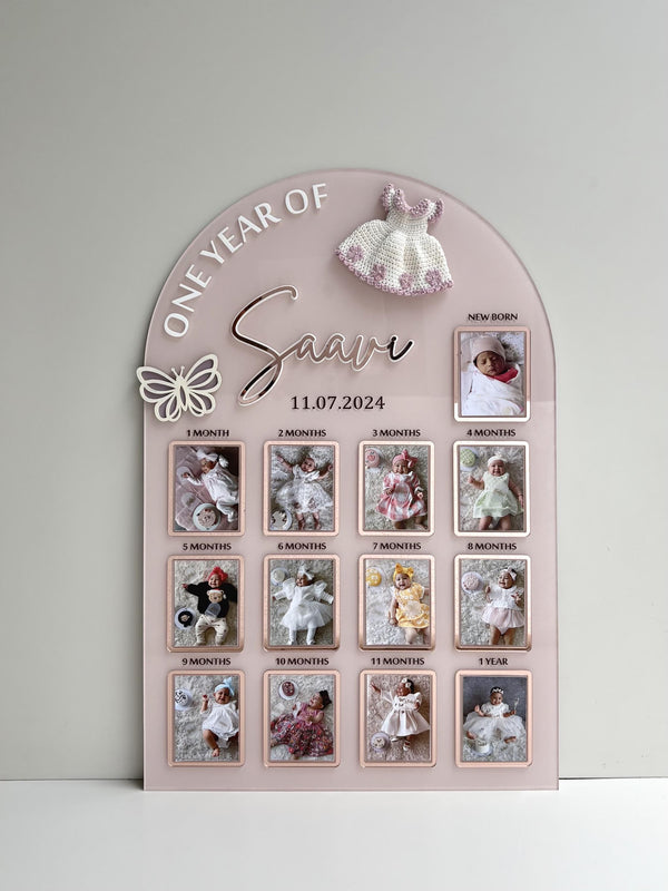 Arch-shaped pink acrylic milestone board with thirteen photo frames from newborn to one year and a crocheted dress topper