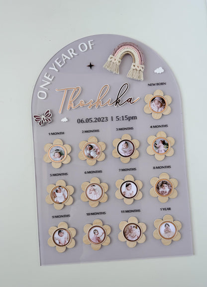Arch-shaped acrylic milestone board with thirteen flower-shaped photo frames from newborn to one year, a macrame rainbow topper, and the baby's name and birth date