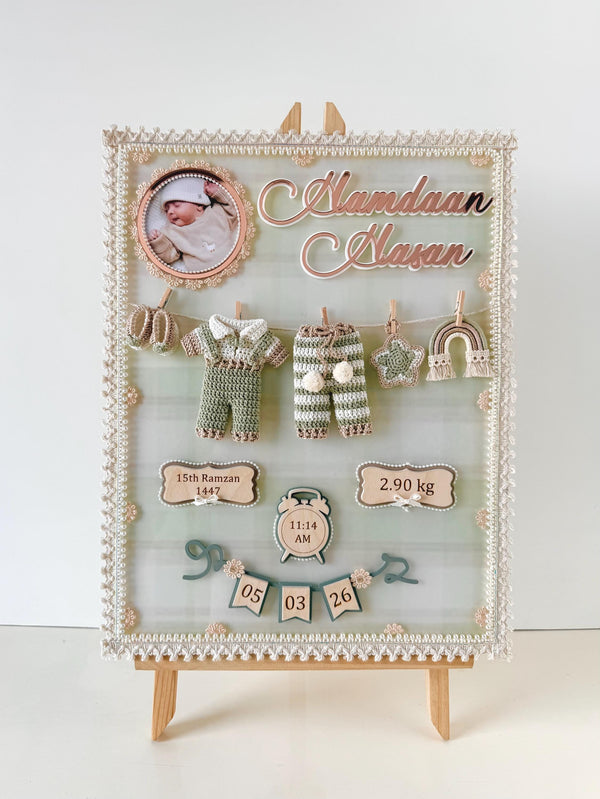 Personalized birth-details keepsake board with a circular baby photo, a tiny crocheted onesie and pants clipped to a mini clothesline, and wooden birth-date, weight, and time tags