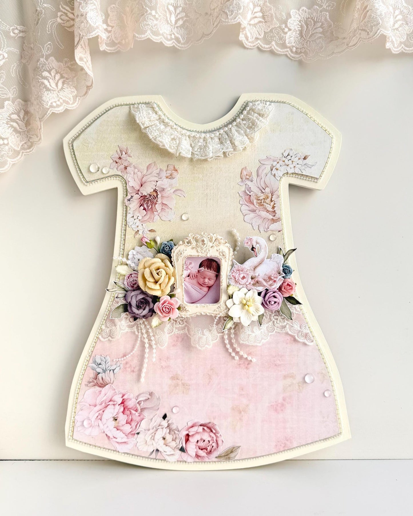 Dress-shaped wooden keepsake frame with pink paper florals, lace trim, an oval baby photo, and a paper swan accent