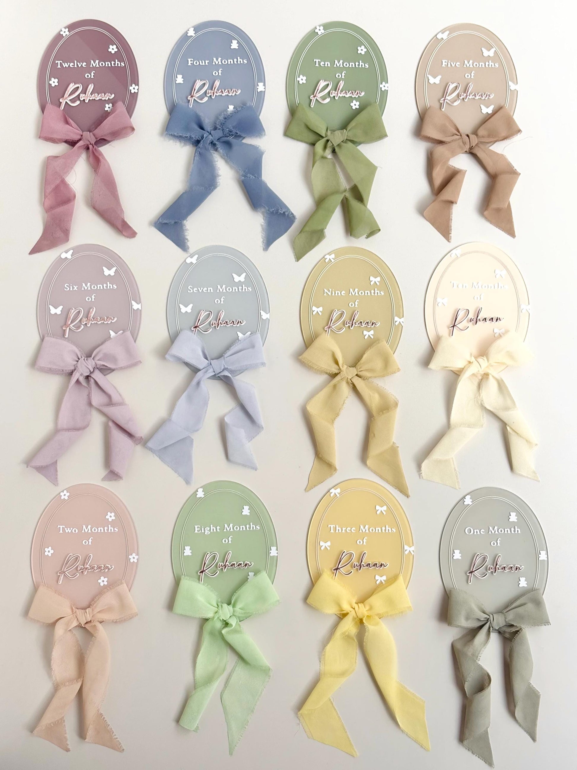 Set of twelve pastel oval acrylic monthly milestone discs, each personalized with the baby's name and a matching fabric bow