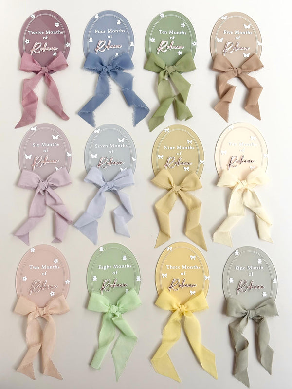 Set of twelve pastel oval acrylic monthly milestone discs, each personalized with the baby's name and a matching fabric bow